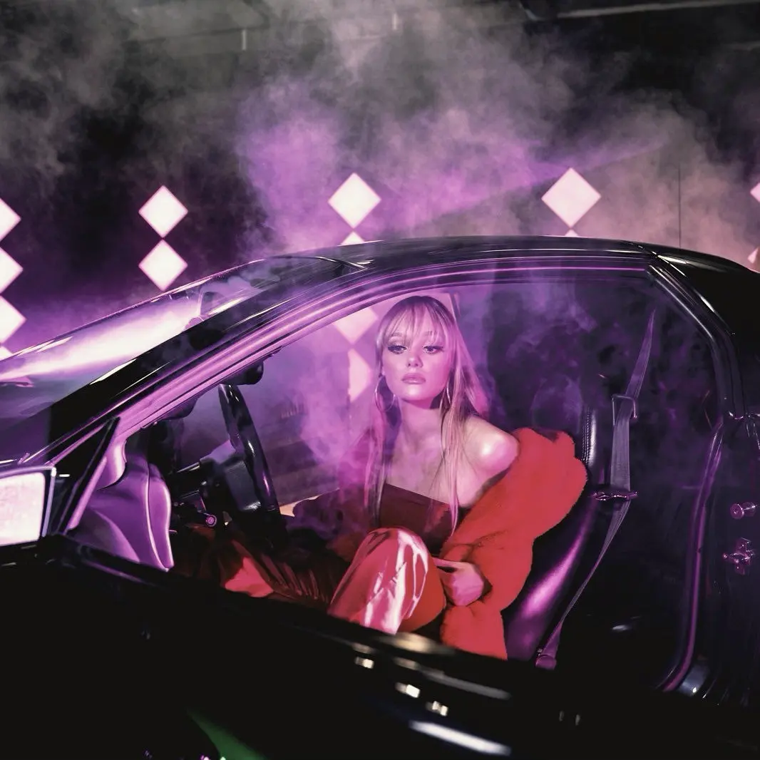 Singer in a red jacket sitting in a car in purple smoke
